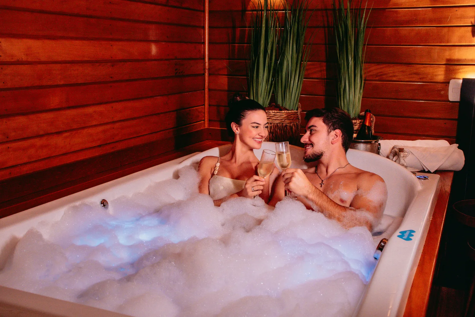 Casal na banheira de hidromassagem da suíte, com taças e luz baixa