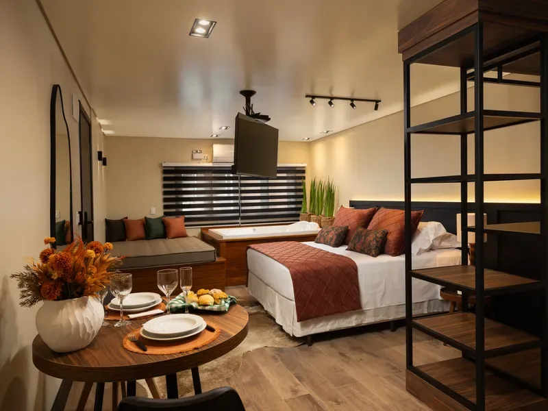Mesa redonda posta na suíte com dois pratos, taças, arranjo de flores secas e fruteira, tendo ao fundo a cama, o day bed junto à janela e a banheira de hidromassagem